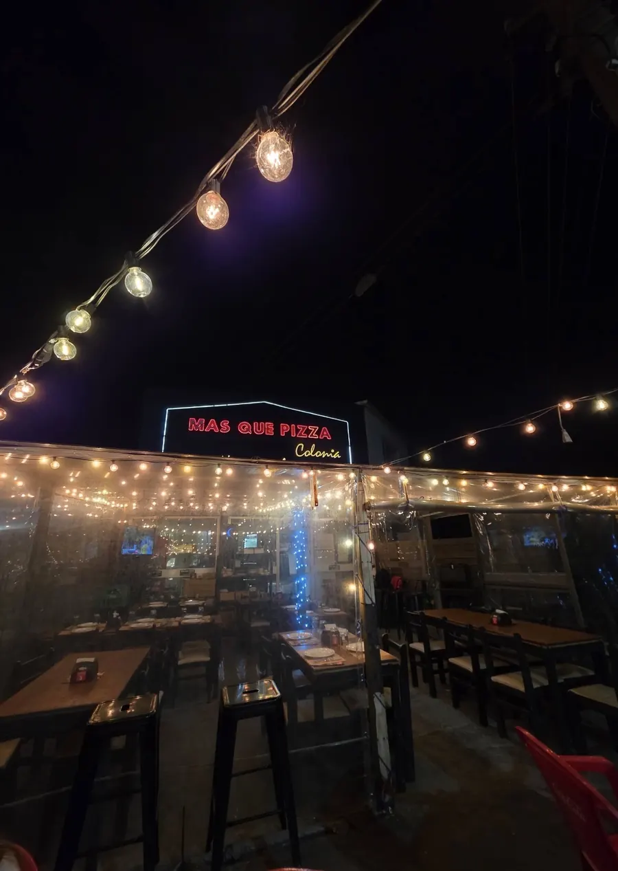 Fachada de Más Que Pizza Colonia de noche, con guirnaldas de luces, mesas de madera y el cartel de neón rojo